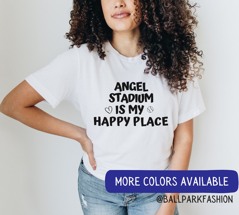 May include: A white t-shirt with black text that reads "Angel Stadium is my happy place" with a heart and baseball graphic. The text is in a handwritten font. The model is wearing the shirt and has long curly brown hair. The text "More colors available @ballparkfashion" is at the bottom of the image.