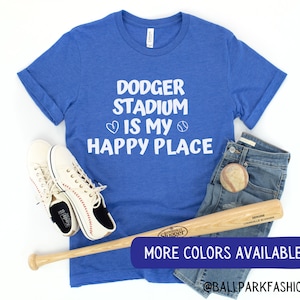 May include: A blue t-shirt with the text "Dodger Stadium is my happy place" in white lettering with a heart and baseball graphic. The shirt is laid out on a white surface with a baseball bat, baseball, and white sneakers. The text "More Colors Available" is in a blue rectangle at the bottom of the image. The text "@ballparkfashion" is below the rectangle.