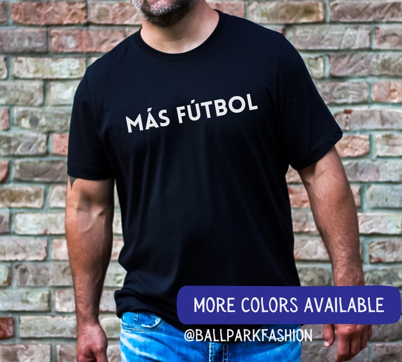 Puede incluir: Una camiseta negra con texto blanco que dice "M&Aacute;s F&Uacute;TBOL". El texto est&aacute; centrado en la camiseta. La camiseta la lleva puesta una persona. La imagen tambi&eacute;n incluye el texto "MORE COLORS AVAILABLE" y "@BALLPARKFASHION".