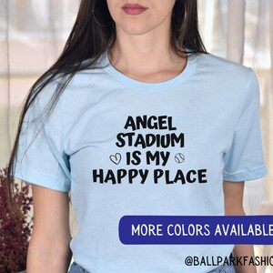 May include: A light blue t-shirt with black text that reads "Angel Stadium is my happy place" with a heart and baseball graphic. The text "More Colors Available" is on a blue rectangle at the bottom of the image. The text "@ballparkfashion" is below the rectangle.
