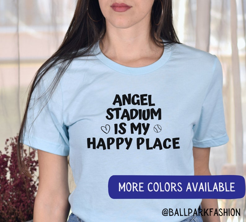 May include: A light blue t-shirt with black text that reads "Angel Stadium is my happy place" with a heart and baseball graphic. The text "More Colors Available" is on a blue rectangle at the bottom of the image. The text "@ballparkfashion" is below the rectangle.