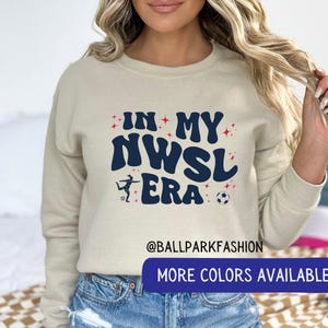 Op de afbeelding: Beige sweatshirt met de tekst "IN MY NWSL ERA" in marineblauw, met een voetbal en een speler afbeelding. De sweatshirt heeft ook de tekst "MORE COLORS AVAILABLE" op een marineblauwe banner.