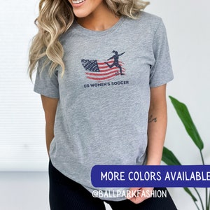 Puede incluir: Camiseta gris con un dise&ntilde;o de bandera estadounidense desgastado y una silueta de un jugador de f&uacute;tbol pateando una pelota. El texto "US Women's Soccer" est&aacute; impreso debajo del dise&ntilde;o. La camiseta est&aacute; siendo usada por una persona. El texto "More Colors Available @BallparkFashion" est&aacute; impreso en una pancarta azul en la parte inferior de la imagen.