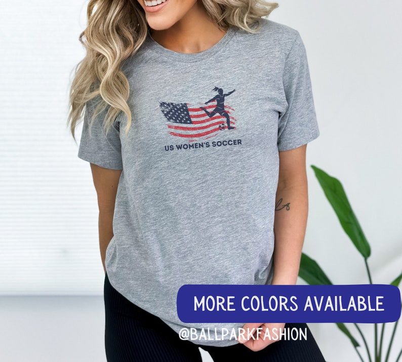 Puede incluir: Camiseta gris con un dise&ntilde;o de bandera estadounidense desgastado y una silueta de un jugador de f&uacute;tbol pateando una pelota. El texto "US Women's Soccer" est&aacute; impreso debajo del dise&ntilde;o. La camiseta est&aacute; siendo usada por una persona. El texto "More Colors Available @BallparkFashion" est&aacute; impreso en una pancarta azul en la parte inferior de la imagen.