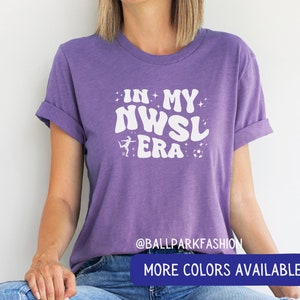 Pu&ograve; includere: Maglietta viola con testo bianco che recita "In My NWSL Era" con una grafica di un calciatore e un pallone da calcio. Il testo &egrave; in uno stile di font retr&ograve;. La maglietta ha un collo rotondo e maniche corte.  @ballparkfashion Altri colori disponibili