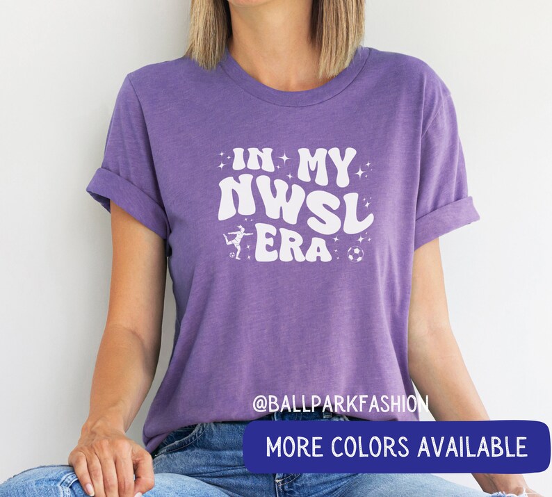 Pu&ograve; includere: Maglietta viola con testo bianco che recita "In My NWSL Era" con una grafica di un calciatore e un pallone da calcio. Il testo &egrave; in uno stile di font retr&ograve;. La maglietta ha un collo rotondo e maniche corte.  @ballparkfashion Altri colori disponibili