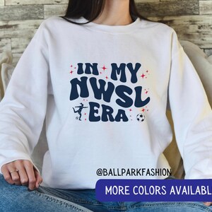 Op de afbeelding: Witte sweatshirt met de tekst "IN MY NWSL ERA" in marineblauw, met een voetbal en een speler grafiek. Rode sterren omringen de tekst. De onderkant van de trui heeft een blauwe banner met "MORE COLORS AVAILABLE".
