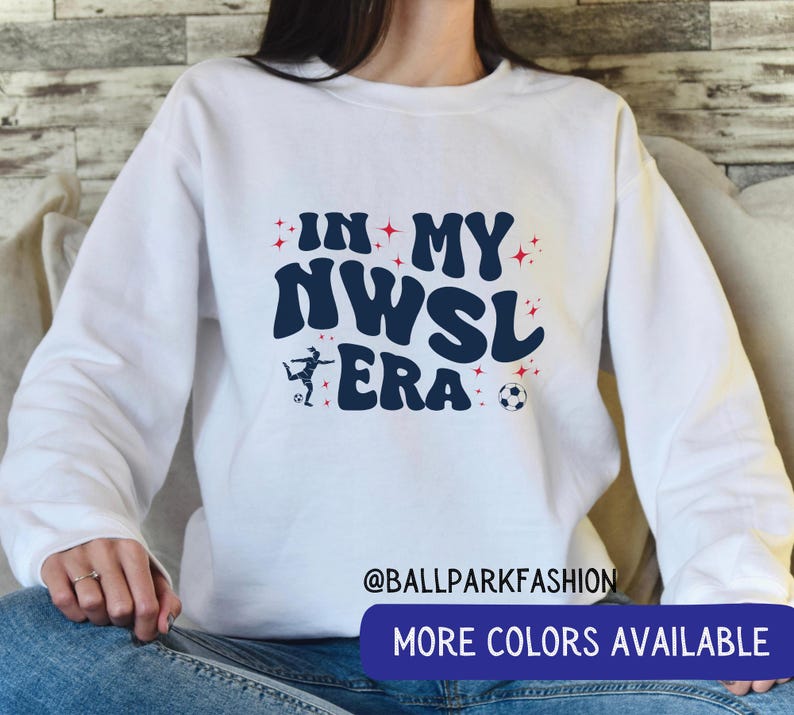 Op de afbeelding: Witte sweatshirt met de tekst "IN MY NWSL ERA" in marineblauw, met een voetbal en een speler grafiek. Rode sterren omringen de tekst. De onderkant van de trui heeft een blauwe banner met "MORE COLORS AVAILABLE".