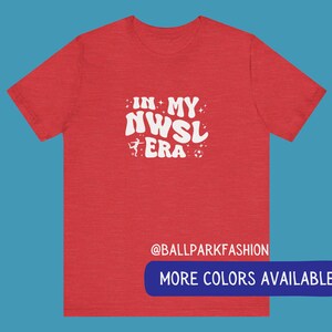 Pu&ograve; includere: T-shirt rosso melange con testo bianco che recita "In My NWSL Era" con un pallone da calcio e una silhouette di un calciatore. Il testo &egrave; in un font retr&ograve;. La parte inferiore della maglietta ha il testo "@ballparkfashion" e un pulsante che dice "Altri colori disponibili".