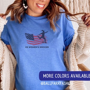 Puede incluir: Una camiseta azul con un gr&aacute;fico de una jugadora de f&uacute;tbol femenino pateando un bal&oacute;n de f&uacute;tbol frente a una bandera estadounidense. El texto "US Women's Soccer" est&aacute; impreso debajo del gr&aacute;fico. La camiseta la lleva puesta una persona con el pelo largo y casta&ntilde;o. El texto "MORE COLORS AVAILABLE @BALLPARKFASHION" est&aacute; impreso en la parte inferior de la imagen.
