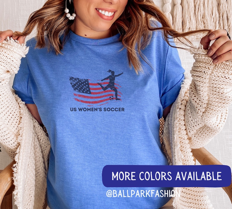 Puede incluir: Una camiseta azul con un gr&aacute;fico de una jugadora de f&uacute;tbol femenino pateando un bal&oacute;n de f&uacute;tbol frente a una bandera estadounidense. El texto "US Women's Soccer" est&aacute; impreso debajo del gr&aacute;fico. La camiseta la lleva puesta una persona con el pelo largo y casta&ntilde;o. El texto "MORE COLORS AVAILABLE @BALLPARKFASHION" est&aacute; impreso en la parte inferior de la imagen.