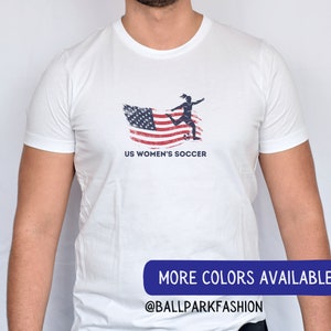 Puede incluir: Camiseta blanca con un gr&aacute;fico de una jugadora de f&uacute;tbol femenino pateando un bal&oacute;n frente a una bandera estadounidense desgastada. El texto "US Women's Soccer" est&aacute; impreso debajo del gr&aacute;fico. El texto "MORE COLORS AVAILABLE @BALLPARKFASHION" est&aacute; impreso en la parte inferior de la imagen.
