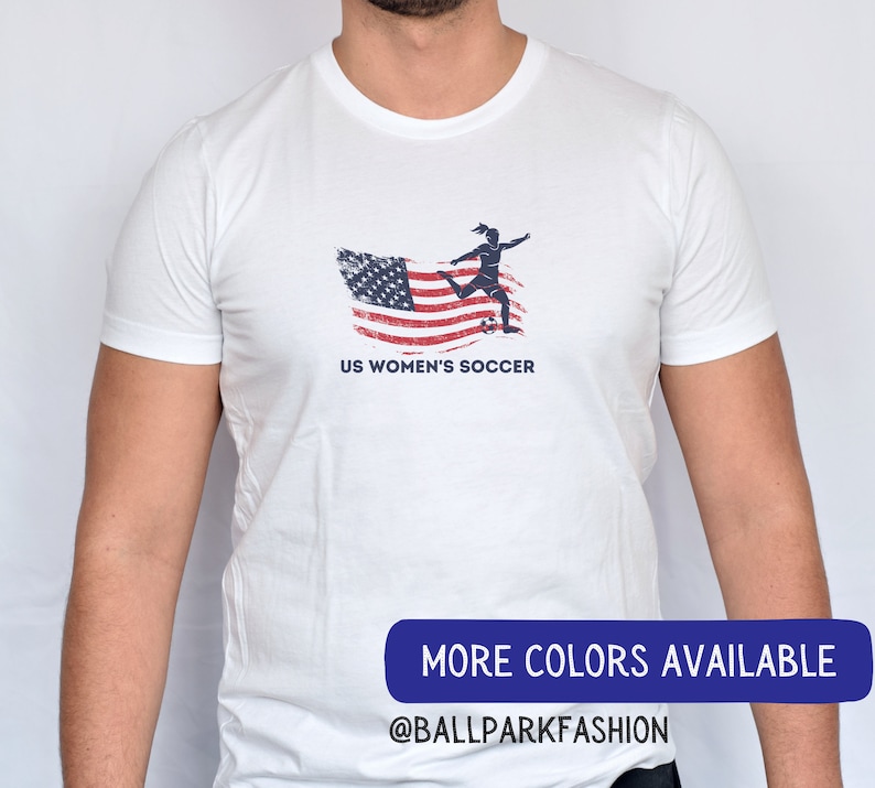 Puede incluir: Camiseta blanca con un gr&aacute;fico de una jugadora de f&uacute;tbol femenino pateando un bal&oacute;n frente a una bandera estadounidense desgastada. El texto "US Women's Soccer" est&aacute; impreso debajo del gr&aacute;fico. El texto "MORE COLORS AVAILABLE @BALLPARKFASHION" est&aacute; impreso en la parte inferior de la imagen.