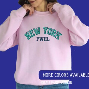 May include: Pink sweatshirt with a teal blue and navy blue graphic that says "NEW YORK PWHL" on the front.