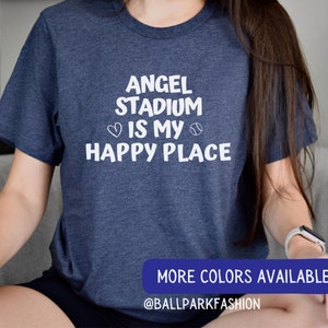 May include: A navy blue t-shirt with white text that reads "Angel Stadium is my happy place" with a heart and baseball graphic. The text "More Colors Available" and "@ballparkfashion" are also visible.