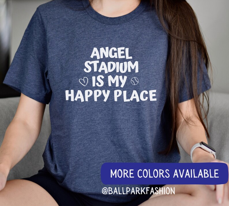 May include: A navy blue t-shirt with white text that reads "Angel Stadium is my happy place" with a heart and baseball graphic. The text "More Colors Available" and "@ballparkfashion" are also visible.