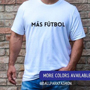 Puede incluir: Camiseta blanca con el texto "M&Aacute;S F&Uacute;TBOL" en letras negras. El texto est&aacute; centrado en la camiseta. La camiseta est&aacute; siendo usada por una persona. La imagen tambi&eacute;n incluye el texto "MORE COLORS AVAILABLE" y "@BALLPARKFASHION" en letras azules.