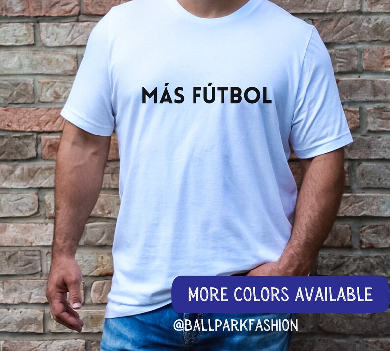 Puede incluir: Camiseta blanca con el texto "M&Aacute;S F&Uacute;TBOL" en letras negras. El texto est&aacute; centrado en la camiseta. La camiseta est&aacute; siendo usada por una persona. La imagen tambi&eacute;n incluye el texto "MORE COLORS AVAILABLE" y "@BALLPARKFASHION" en letras azules.