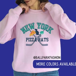 May include: Pink sweatshirt with a graphic design of a rat eating pizza and a hockey puck with the text "New York Pizza Rats" and the text "@ballparkfashion" and "More Colors Available" below the graphic.