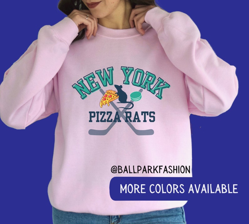 May include: Pink sweatshirt with a graphic design of a rat eating pizza and a hockey puck with the text "New York Pizza Rats" and the text "@ballparkfashion" and "More Colors Available" below the graphic.