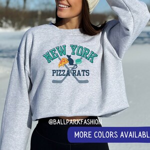May include: A grey cropped sweatshirt with a graphic of a rat eating pizza and a hockey puck with the text "New York Pizza Rats" in teal and white lettering. The text "@ballparkfashion" is at the bottom of the sweatshirt.