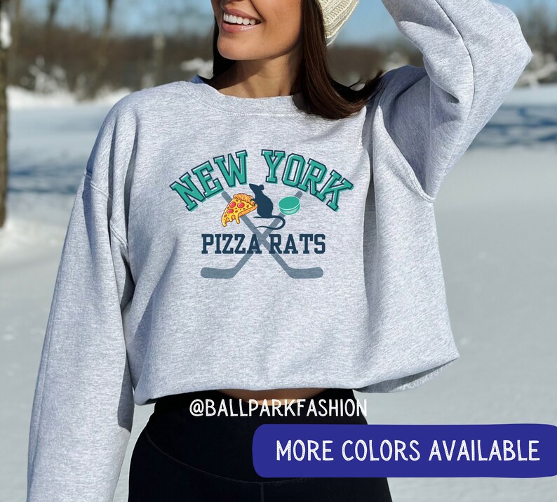 May include: A grey cropped sweatshirt with a graphic of a rat eating pizza and a hockey puck with the text "New York Pizza Rats" in teal and white lettering. The text "@ballparkfashion" is at the bottom of the sweatshirt.