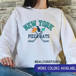 May include: White sweatshirt with a graphic design featuring the words "New York Pizza Rats" in teal and black lettering. The graphic also includes a pizza slice, a rat, and two hockey sticks.