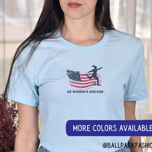Puede incluir: Camiseta azul claro con un gr&aacute;fico de una jugadora de f&uacute;tbol femenino pateando un bal&oacute;n de f&uacute;tbol frente a una bandera estadounidense desgastada. El texto "US Women's Soccer" est&aacute; impreso debajo del gr&aacute;fico. El texto "More Colors Available @BallparkFashion" est&aacute; impreso en la parte inferior de la imagen.