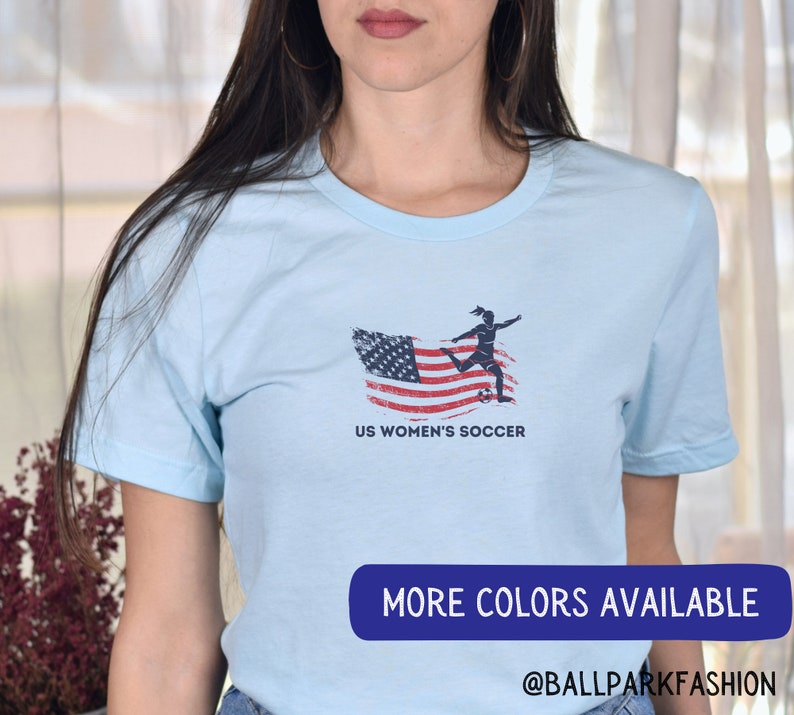 Puede incluir: Camiseta azul claro con un gr&aacute;fico de una jugadora de f&uacute;tbol femenino pateando un bal&oacute;n de f&uacute;tbol frente a una bandera estadounidense desgastada. El texto "US Women's Soccer" est&aacute; impreso debajo del gr&aacute;fico. El texto "More Colors Available @BallparkFashion" est&aacute; impreso en la parte inferior de la imagen.