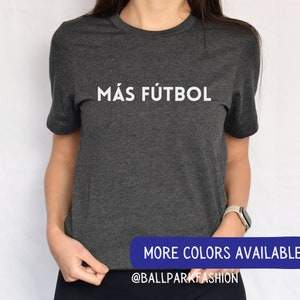 Puede incluir: Una camiseta gris oscuro con el texto "M&Aacute;S F&Uacute;TBOL" impreso en blanco en la parte delantera. El texto est&aacute; centrado en la camiseta. La camiseta est&aacute; hecha de un material suave y c&oacute;modo y es perfecta para el uso diario.