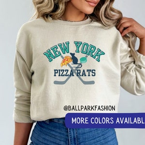 May include: A light beige sweatshirt with a graphic design featuring the words "New York Pizza Rats" in teal and white. The graphic also includes a cartoon rat holding a slice of pizza and a hockey stick. The text "@ballparkfashion" is printed below the graphic. The sweatshirt has long sleeves and a crew neck.