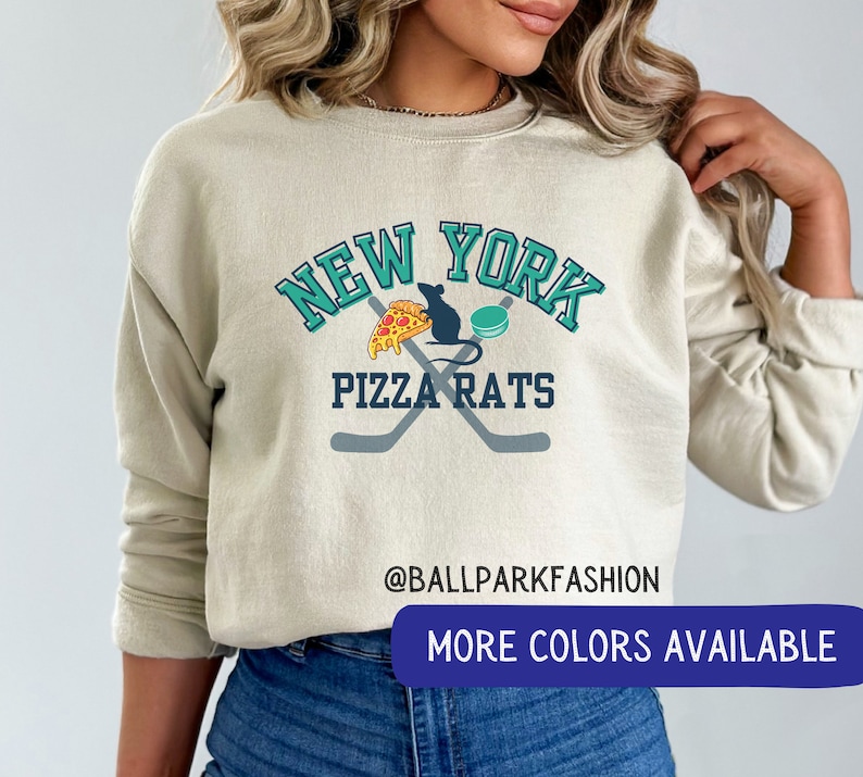May include: A light beige sweatshirt with a graphic design featuring the words "New York Pizza Rats" in teal and white. The graphic also includes a cartoon rat holding a slice of pizza and a hockey stick. The text "@ballparkfashion" is printed below the graphic. The sweatshirt has long sleeves and a crew neck.