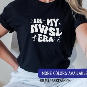 Pu&ograve; includere: Una maglietta nera con testo bianco che recita "In My NWSL Era" con un calciatore stilizzato e un'immagine di un pallone da calcio. Il testo &egrave; in un font retr&ograve;. La maglietta &egrave; indossata da una persona. Il testo "More Colors Available @BALLPARKFASHION" &egrave; in basso nell'immagine.