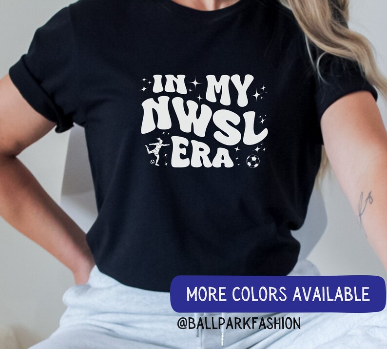 Pu&ograve; includere: Una maglietta nera con testo bianco che recita "In My NWSL Era" con un calciatore stilizzato e un'immagine di un pallone da calcio. Il testo &egrave; in un font retr&ograve;. La maglietta &egrave; indossata da una persona. Il testo "More Colors Available @BALLPARKFASHION" &egrave; in basso nell'immagine.