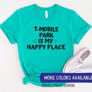 May include: A turquoise t-shirt with the text "T-Mobile Park is my happy place" in black lettering. The text is surrounded by a heart and a baseball graphic. The shirt is tied in a knot at the bottom. The text "MORE COLORS AVAILABLE @BALLPARKFASHION" is at the bottom of the image.