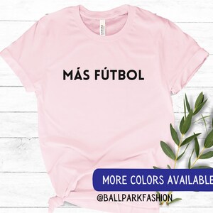 Puede incluir: Camiseta rosa con el texto "M&Aacute;S F&Uacute;TBOL" en letras negras. El texto est&aacute; centrado en la camiseta. La camiseta est&aacute; sobre un fondo de madera blanca. El texto "MORE COLORS AVAILABLE" y "@BALLPARKFASHION" son visibles en la parte inferior de la imagen.