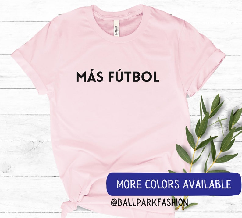 Puede incluir: Camiseta rosa con el texto "M&Aacute;S F&Uacute;TBOL" en letras negras. El texto est&aacute; centrado en la camiseta. La camiseta est&aacute; sobre un fondo de madera blanca. El texto "MORE COLORS AVAILABLE" y "@BALLPARKFASHION" son visibles en la parte inferior de la imagen.