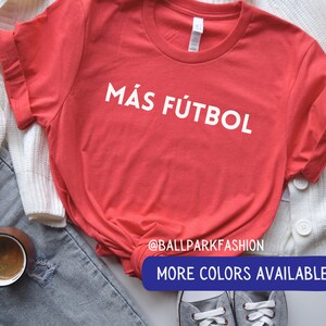 Puede incluir: Camiseta roja con texto blanco que dice "M&aacute;s F&uacute;tbol". La camiseta est&aacute; puesta sobre una superficie blanca con un par de zapatillas grises y un par de pantalones vaqueros azules en el fondo. El texto "@ballparkfashion" y "M&aacute;s colores disponibles" tambi&eacute;n es visible en la imagen.