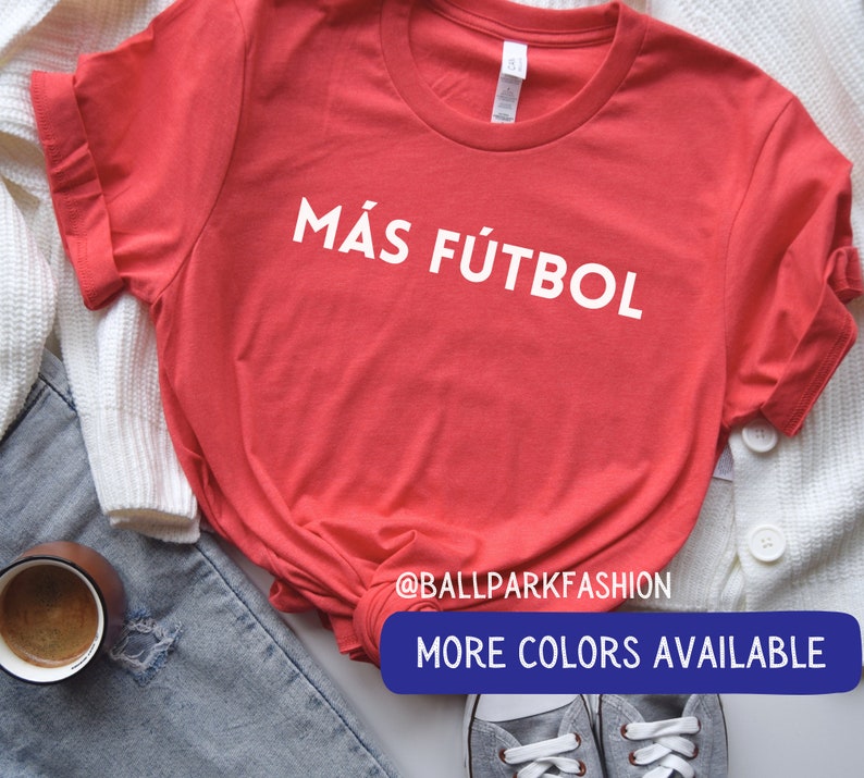 Puede incluir: Camiseta roja con texto blanco que dice "M&aacute;s F&uacute;tbol". La camiseta est&aacute; puesta sobre una superficie blanca con un par de zapatillas grises y un par de pantalones vaqueros azules en el fondo. El texto "@ballparkfashion" y "M&aacute;s colores disponibles" tambi&eacute;n es visible en la imagen.