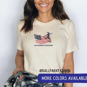Puede incluir: Una camiseta blanca con un dise&ntilde;o de bandera estadounidense desgastada y una silueta de un jugador de f&uacute;tbol pateando una pelota. El texto "US Women's Soccer" est&aacute; impreso debajo de la imagen. La camiseta tiene cuello redondo y mangas cortas.  @ballparkfashion M&Aacute;S COLORES DISPONIBLES