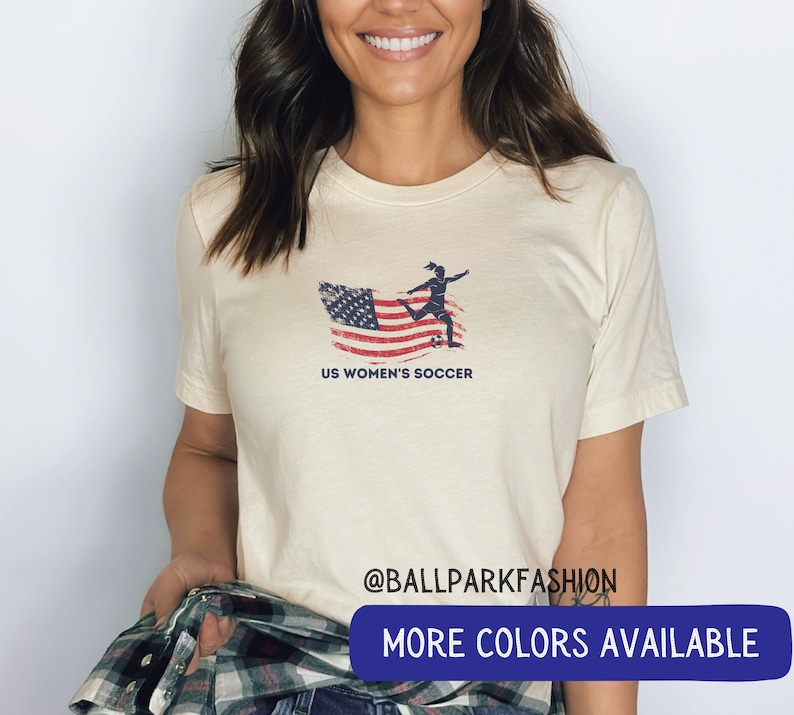 Puede incluir: Una camiseta blanca con un dise&ntilde;o de bandera estadounidense desgastada y una silueta de un jugador de f&uacute;tbol pateando una pelota. El texto "US Women's Soccer" est&aacute; impreso debajo de la imagen. La camiseta tiene cuello redondo y mangas cortas.  @ballparkfashion M&Aacute;S COLORES DISPONIBLES