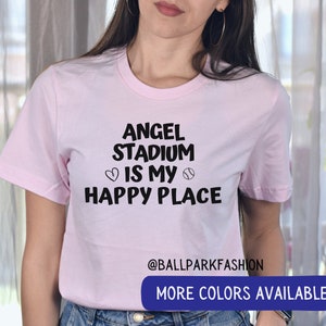 May include: Pink t-shirt with the text "Angel Stadium is my happy place" in black lettering with a heart and baseball graphic. The text "@ballparkfashion" is at the bottom of the shirt. The text "More Colors Available" is in a blue box at the bottom of the image.