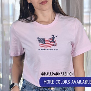 Puede incluir: Camiseta rosa con un gr&aacute;fico de bandera estadounidense desgastado y una silueta de una mujer pateando un bal&oacute;n de f&uacute;tbol. El texto "US Women's Soccer" est&aacute; impreso debajo del gr&aacute;fico. El texto "@ballparkfashion" est&aacute; impreso en la parte inferior de la camiseta. El texto "M&aacute;s colores disponibles" est&aacute; impreso en un rect&aacute;ngulo azul en la parte inferior de la imagen.