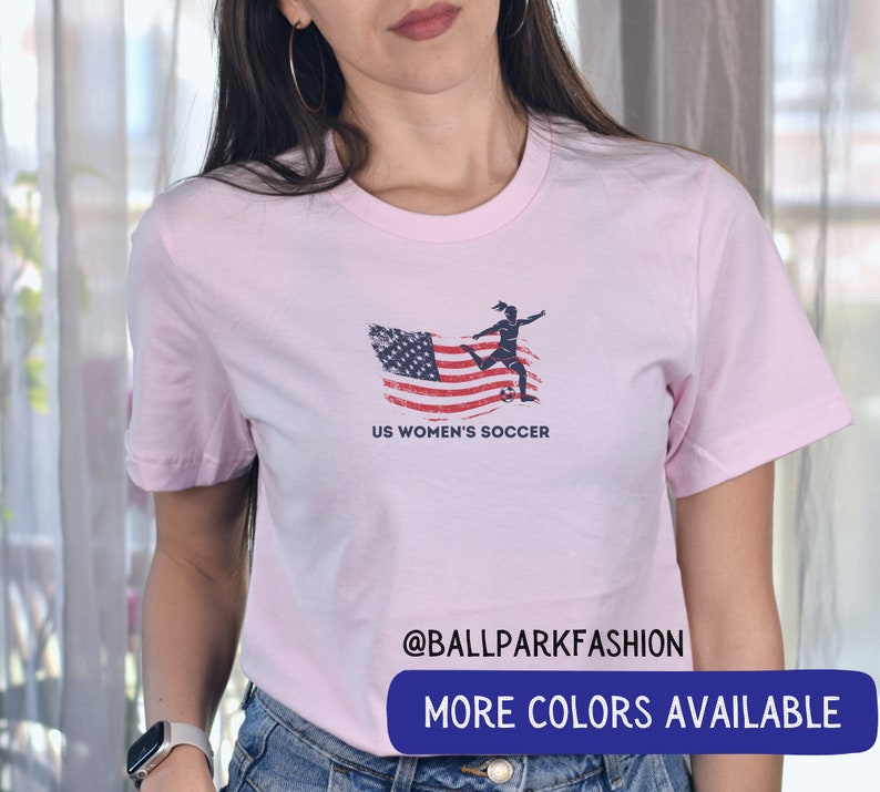 Puede incluir: Camiseta rosa con un gr&aacute;fico de bandera estadounidense desgastado y una silueta de una mujer pateando un bal&oacute;n de f&uacute;tbol. El texto "US Women's Soccer" est&aacute; impreso debajo del gr&aacute;fico. El texto "@ballparkfashion" est&aacute; impreso en la parte inferior de la camiseta. El texto "M&aacute;s colores disponibles" est&aacute; impreso en un rect&aacute;ngulo azul en la parte inferior de la imagen.