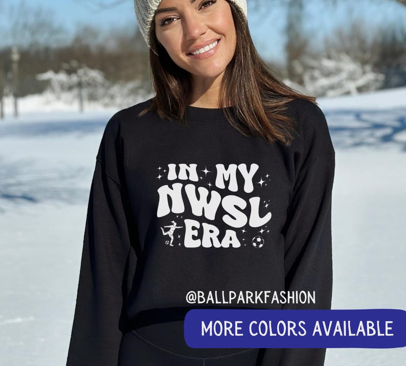Op de afbeelding: Zwart sweatshirt met witte tekst "IN MY NWSL ERA", met een voetbal en een speler. De tekst "@BALLPARKFASHION" staat eronder, met een knop "MORE COLORS AVAILABLE".