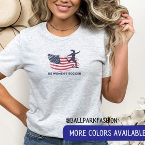 Puede incluir: Una camiseta gris jaspeada con un gr&aacute;fico de una jugadora de f&uacute;tbol pateando una pelota frente a una bandera estadounidense. El texto "US Women's Soccer" est&aacute; impreso debajo del gr&aacute;fico. La camiseta la lleva puesta una persona con el pelo largo y rubio. El texto "@ballparkfashion" y "M&aacute;s colores disponibles" est&aacute; impreso debajo de la imagen.