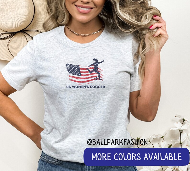 Puede incluir: Una camiseta gris jaspeada con un gr&aacute;fico de una jugadora de f&uacute;tbol pateando una pelota frente a una bandera estadounidense. El texto "US Women's Soccer" est&aacute; impreso debajo del gr&aacute;fico. La camiseta la lleva puesta una persona con el pelo largo y rubio. El texto "@ballparkfashion" y "M&aacute;s colores disponibles" est&aacute; impreso debajo de la imagen.