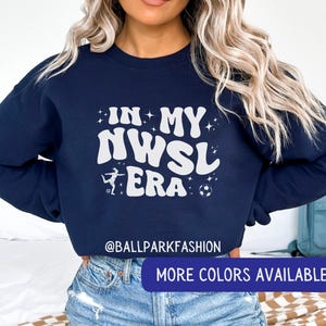 Op de afbeelding: Marineblauw sweatshirt met de witte tekst "IN MY NWSL ERA" en voetbalthema graphics. Het sweatshirt heeft ook de tekst "@BALLPARKFASHION" en een knop met de tekst "MORE COLORS AVAILABLE".