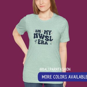 Pu&ograve; includere: Una maglietta blu chiaro con il testo "In My NWSL Era" in blu navy con una stella rossa e un'immagine di pallone da calcio. Il testo &egrave; in un font retr&ograve;. La maglietta ha un collo rotondo e maniche corte. Il testo "@ballparkfashion" &egrave; in fondo alla maglietta. Il testo "Altri colori disponibili" &egrave; in un rettangolo blu in fondo all'immagine.