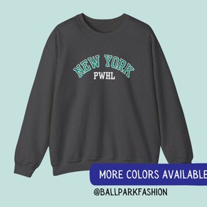 May include: A dark grey sweatshirt with a green and white "NEW YORK" graphic with the letters "PWHL" underneath. The text is in a collegiate style font. The sweatshirt has long sleeves and a crew neck.